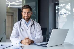 retrato do uma sorridente jovem masculino médico dentro uma branco casaco sentado dentro a escritório do a clínica às a trabalhando mesa ,e com confiança olhando às a Câmera. foto