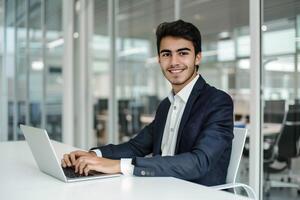 uma jovem homem dentro uma terno sentado às uma branco escrivaninha com uma computador portátil dentro escritório foto