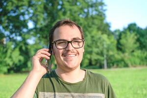jovem homem com Óculos e verde camiseta falando em célula telefone às a parque foto