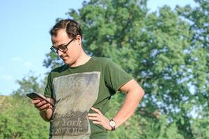 jovem homem com Óculos e verde camiseta mensagens de texto em célula telefone às parque foto