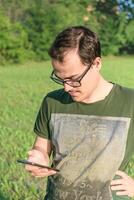 jovem homem com Óculos e verde camiseta mensagens de texto em célula telefone às parque foto