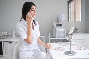 fêmea cosmetologista falando em Smartphone às escrivaninha dentro beleza clínica foto