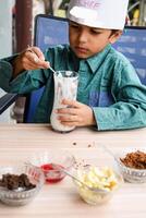 fofa indiano chefe de cozinha Garoto preparando sundae prato Como uma parte do não fogo cozinhando que inclui baunilha gelo creme, Brownie, coco pó, recentemente picado frutas e morango xarope. pequeno criança preparando Comida foto