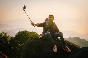 homem é levando uma selfie com dele célula telefone enquanto sentado em uma Rocha foto