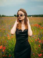 lindo jovem menina dentro uma Preto tarde vestir e oculos de sol posando contra uma papoula campo em uma nublado verão dia. retrato do uma fêmea modelo ao ar livre. chuvoso clima. cinzento nuvens. foto