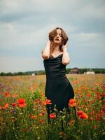 lindo jovem menina dentro uma Preto tarde vestir posando contra uma papoula campo em uma nublado verão dia. retrato do uma fêmea modelo ao ar livre. chuvoso clima. cinzento nuvens. foto