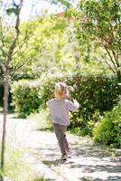 pequeno menina corre ao longo uma caminho dentro uma verde parque, acenando dela braços. costas Visão foto