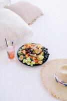 coquetel com uma Palha dentro uma vidro e uma prato do frutas ficar de pé Próximo para uma chapéu em uma branco chaise salão foto