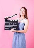 menina adolescente feliz e sorridente em um vestido azul segurando uma claquete isolada em rosa
