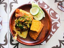 indonésio Comida. frango penyet com Pimenta molho com tofu, tempeh, pepino, ovo e de outros fresco legumes em madeira prato foto