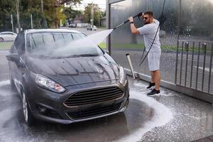 uma homem completamente lavagens a sujeira fora a carro com uma água canhão. uma carro às uma auto serviço carro lavar. foto