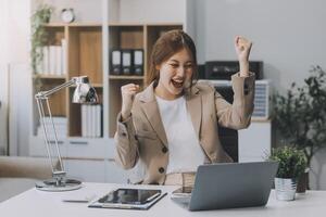 feliz empresária asiática de sucesso animada triunfando com um smartphone de computador portátil no escritório do local de trabalho foto