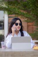 escritório lindo ásia mulher dentro ao ar livre cafeteria. a elegante menina chamadas alguém vestindo oculos de sol para conteúdo ou publicidade foto