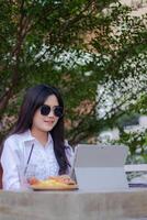 escritório lindo ásia mulher dentro ao ar livre cafeteria. sorridente menina fazendo trabalhos em computador portátil dentro oculos de sol sincero para conteúdo ou publicidade foto