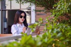 escritório lindo ásia mulher dentro ao ar livre cafeteria. elegante menina verificação trabalhos em computador portátil segurando caneta vestindo oculos de sol para conteúdo ou publicidade foto