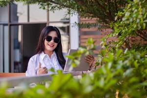 escritório lindo ásia mulher dentro ao ar livre cafeteria. sorridente menina com uma polegares acima placa com sucesso completando uma trabalho em uma computador portátil vestindo oculos de sol para conteúdo ou publicidade foto