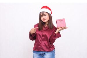 retrato do uma lindo ásia menina dentro dela 20s carregando uma presente caixa às Natal santa claus chapéu vestindo uma moderno brilhante vermelho camisa apontando às a caixa sorridente em uma branco fundo para promoção foto