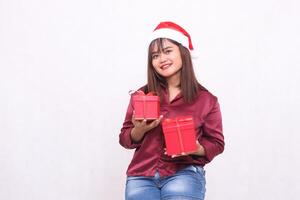 lindo jovem ásia indonésio menina sorridente carregando presente caixa às Natal santa claus chapéu moderno vermelho camisa equipamento elevação caixa acima e baixa em branco fundo para promoção e publicidade foto
