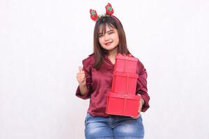 lindo jovem mulher a partir de sudeste Ásia sorrisos com a OK placa enquanto carregando 3 caixas do presentes às Natal vestindo uma santa arco de cabelo e uma moderno vermelho camisa em uma branco fundo para promoção foto