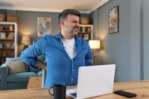 tiro do uma homem de negocios sofrimento a partir de uma dor lombar enquanto trabalhando às dele escrivaninha dentro dele escritório. homem tendo costas dor enquanto trabalhando em computador portátil a partir de casa foto