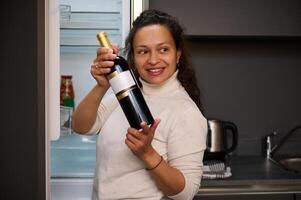 feliz sorridente mulher em pé de frigorífico, segurando uma garrafa do vinho com branco em branco brincar rótulo, escolhendo beber para jantar dentro a casa cozinha. Comida e beber consumismo foto