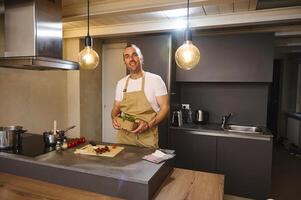 bonito jovem adulto homem anos 40, dentro bege chefe de cozinha avental segurando uma cartão engradado com fresco verde folhas do Rúcula, sorridente olhando às Câmera, cozinhando saudável jantar dentro a casa cozinha. foto