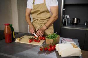 fechar-se Visão do uma masculino chefe de cozinha dentro bege avental, cortar tomate cereja em uma corte borda. ingredientes em a cozinha mesa. homem cozinhando saudável salada dentro a minimalista casa cozinha interior foto