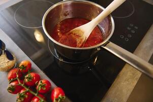 Visão a partir de acima de madeira colher dentro uma aço panela com ebulição caseiro molhos do maduro orgânico suculento vermelho tomates. ramo do fresco vermelho tomate cereja em a cozinha contador perto uma Preto indução fogão foto