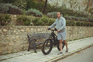 ao ar livre cheio tiro do uma feliz jovem desportivo homem empurrando acima a rua dele elétrico bateria alimentado bicicleta, montanha bicicleta foto