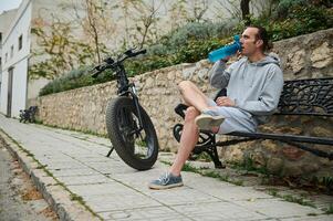cheio comprimento retrato ciclista homem bebendo água, sentado em cidade banco. elétrico moto, bicicleta elétrica, elétrico bicicleta bicicleta em a primeiro plano. bicicleta partilha cidade serviço. sustentabilidade. foto