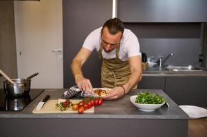 europeu homem dentro bege chef avental, em pé às cozinha contador e usando cozinha toalha, removendo vestígios do tomate molho em uma prato do recentemente preparado italiano massa. pessoas. culinária. italiano cozinha foto