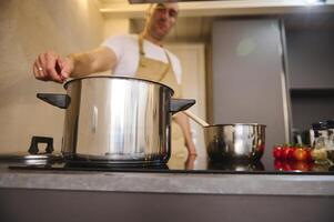 seletivo foco em uma inoxidável aço panela em indução fogão chefe de cozinha preparando Comida dentro a moderno minimalista casa cozinha interior foto