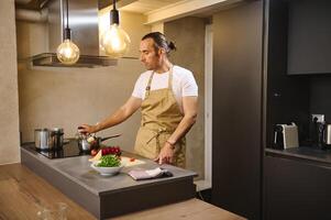 bonito homem cozinhando saudável jantar dentro moderno cozinha interior. atraente cara, chefe de cozinha dentro bege avental, em pé às cozinha contador com fresco orgânico ingredientes, preparando delicioso refeição foto