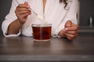 fechar-se uma mulher mãos segurando uma colher com açúcar. fêmea mãos segurando uma colher de chá com açúcar, adoçante a beber dentro uma vidro. adicionando açúcar para uma copo do quente beber bebida foto