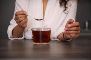 fechar-se mulher mãos segurando uma colher de chá com branco açúcar acima uma vidro copo do recentemente fez Preto chá dentro a casa cozinha, preparando quente beber para café da manhã dentro a manhã foto