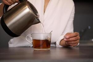 fechar-se do uma mulher fazer chá para café da manhã. detalhes em fêmea mãos derramando ebulição água a partir de uma chaleira para dentro vidro copo com chá bolsa. Comida e beber consumismo foto