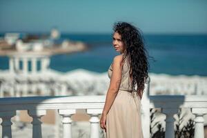 mar mulher descansar. uma mulher com grandes encaracolado cabelo dentro uma bege vestir carrinhos com dela costas e parece às a mar e a costa a partir de uma sacada com balaústres. turista viagem para a mar. foto