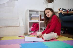 feliz inteligente elementar era escola criança sorridente olhando às Câmera enquanto fazendo dever de casa às acolhedor casa interior foto