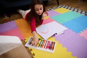 topo Visão do pequeno menina desenhando usando pastel giz de cera enquanto deitado em uma colorida multicolorido enigma tapete em a chão dentro dela quarto às lar. crianças entretenimento, desenvolvimento, arte e Educação conceito foto