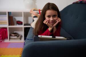 pequeno criança garota, viciado para digital gadgets, jogando conectados jogos enquanto deitado em a sofá às lar. adorável escola menina assistindo filmes, sorridente olhando às Câmera. feliz despreocupado infância foto