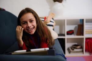 alegre pequeno criança menina sorrisos olhando às Câmera, deitado em sofá às lar, assistindo desenho animado em digital tábua. pequeno criança ter Diversão em moderno gadgets. pessoas. crianças, Internet e digital vício foto