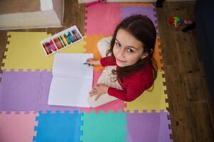 a sobrecarga Visão do uma fofa pequeno criança menina sorridente olhando às Câmera, desenhando com pastel lápis, sentado em uma colorida enigma tapete dentro acolhedor doméstico sala. pessoas. infância. arte e criatividade foto