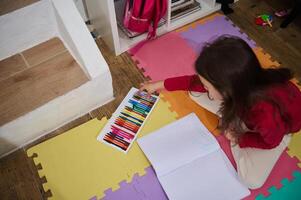 Visão a partir de acima do uma pequeno criança menina levando Fora colorida lápis a partir de uma caixa, desenhando cenário às lar, deitado em uma multi colori enigma lápis foto