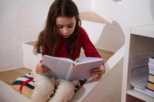 retrato do fofa pequeno escola envelhecido menina focado, segurando livro e lendo texto, sentado em passos às acolhedor casa interior foto