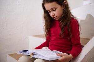 autêntico retrato do inteligente pequeno criança menina folheando através a Páginas do uma capa dura livro, lendo livro, sentado em a passos às lar, sobre branco parede fundo do uma rural casa. cópia de espaço para Publicidades foto