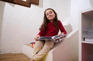 adorável inteligente primário escola aluna menina sorridente olhando às Câmera, sentado em passos às casa com uma livro dentro mãos. inteligente pequeno menina lendo uma livro. mundo livro dia conceito foto