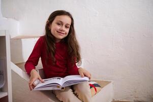 inteligente primário escola aluna menina sorridente às Câmera, sentado em passos às casa com uma livro dentro mãos. pequeno menina lendo uma livro. mundo livro dia conceito. costas para escola em Novo semestre do acadêmico ano foto