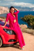 uma mulher dentro uma Rosa terno é sentado em uma vermelho carro. ela é sorridente e posando para a Câmera. a cena tem uma casual e relaxado vibração, com a mulher desfrutando dela Tempo ao ar livre. foto