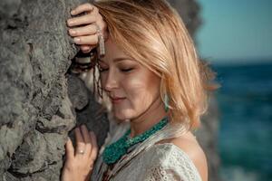 meio envelhecido mulher parece Boa com loiro cabelo, boho estilo dentro branco grandes vestir em a de praia decorações em dela pescoço e braços. foto