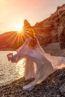 misterioso mulher silhueta grandes cabelo anda em em a de praia oceano água, mar ninfa vento escuta para a aceno. lança acima uma grandes branco vestir, uma divino pôr do sol. artístico foto a partir de a costas sem uma face